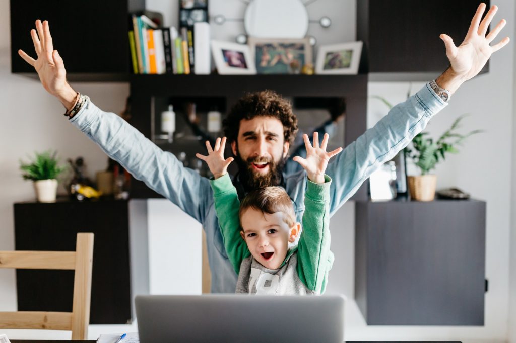 Portrait of happy father and son with arms up sitting at desk with laptop.